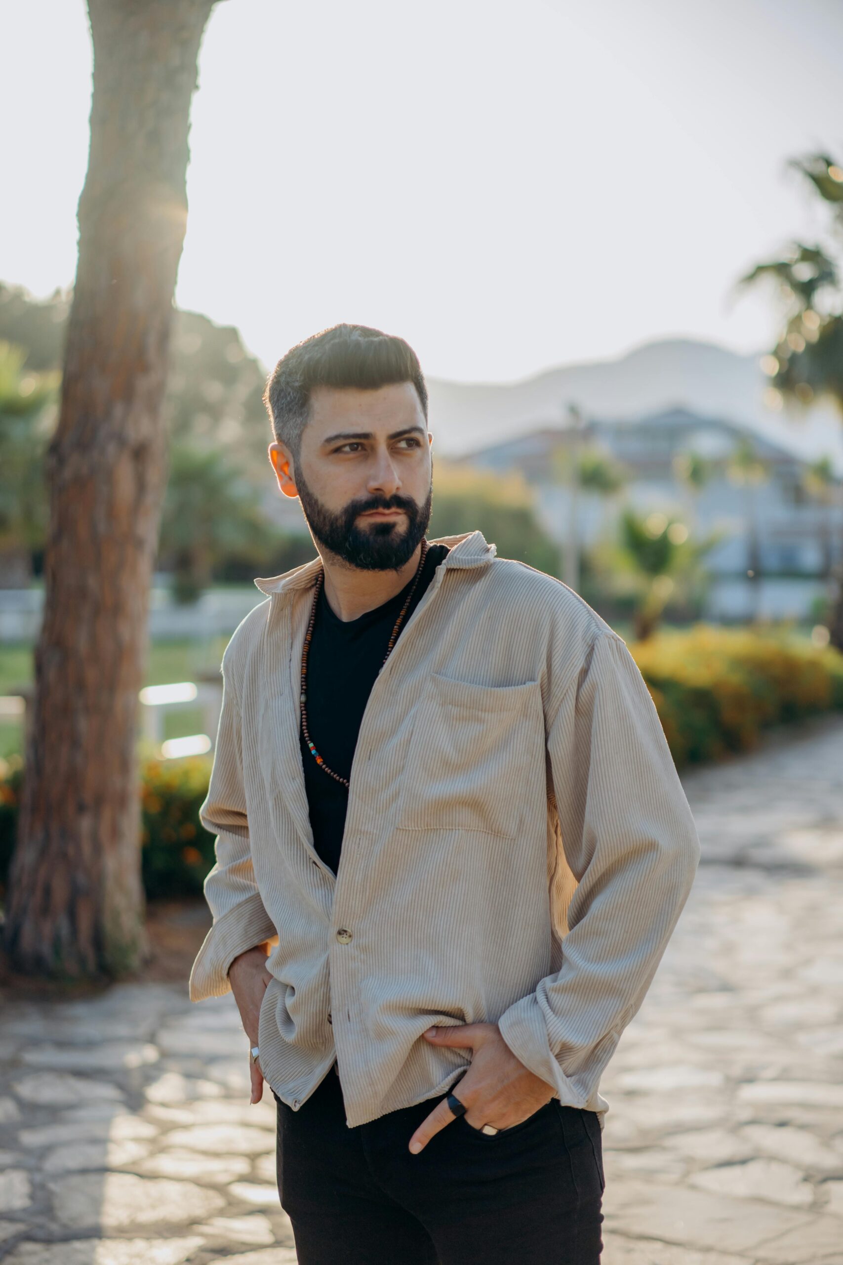Bearded man with hands in pockets posing outdoors. Stylish outfit in natural setting.